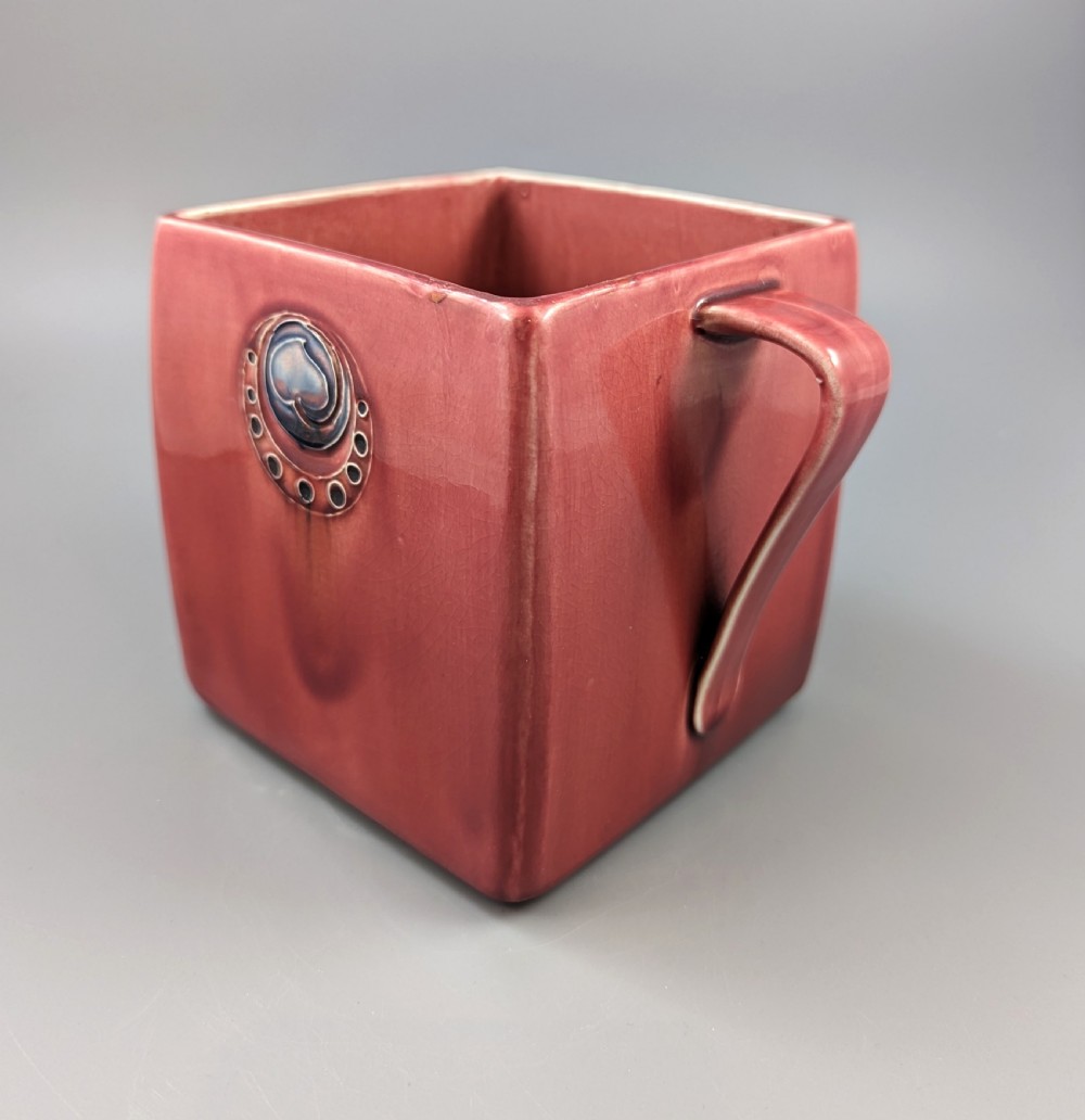 A square red glazed ceramic vessel with twin loop handles and a central circular motif on the front surface. View VII