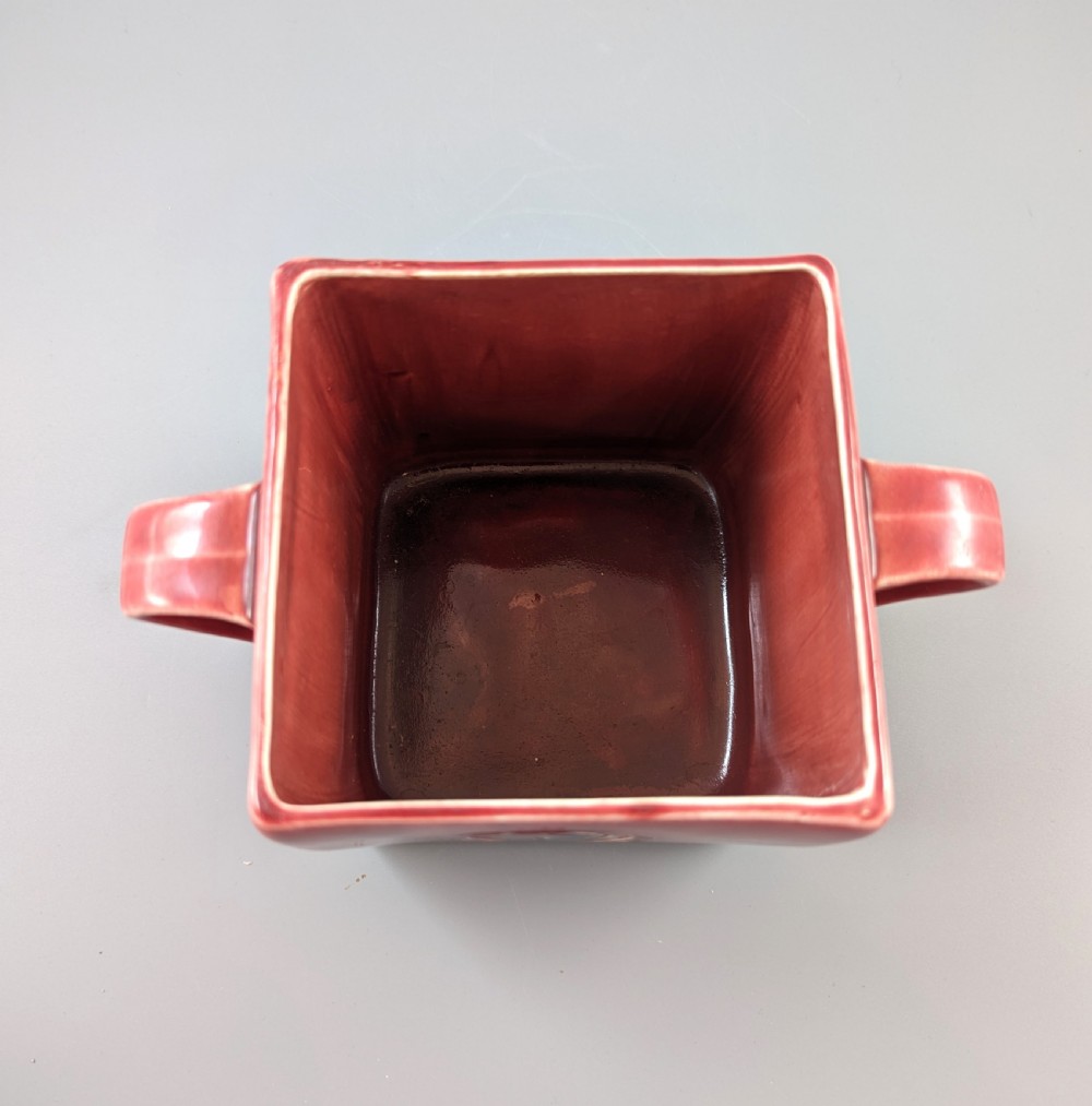 A square red glazed ceramic vessel with twin loop handles and a central circular motif on the front surface. View X