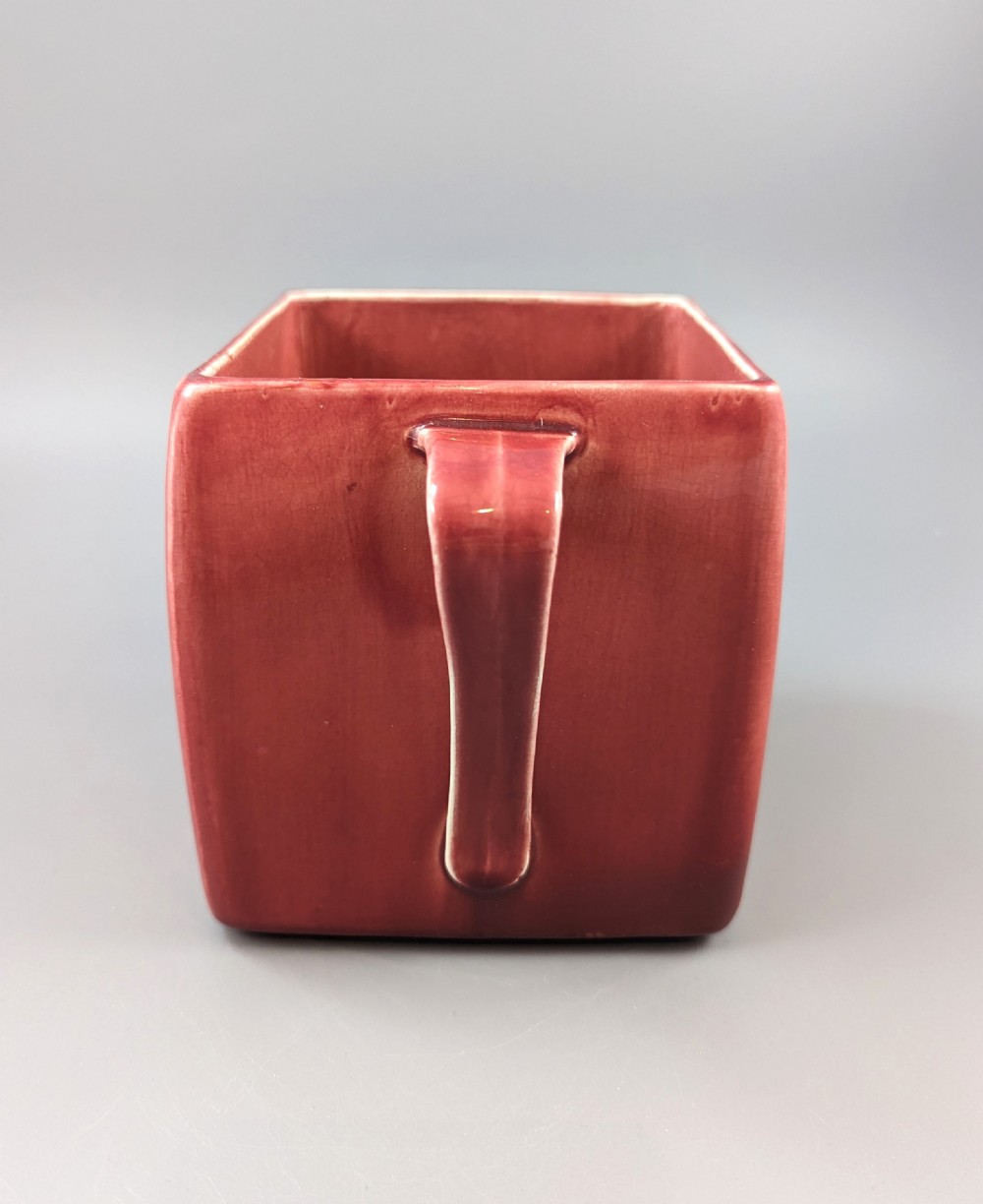 A square red glazed ceramic vessel with twin loop handles and a central circular motif on the front surface. View XI