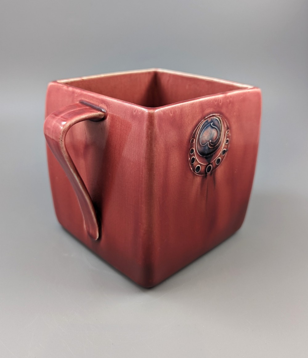 A square red glazed ceramic vessel with twin loop handles and a central circular motif on the front surface. View XII