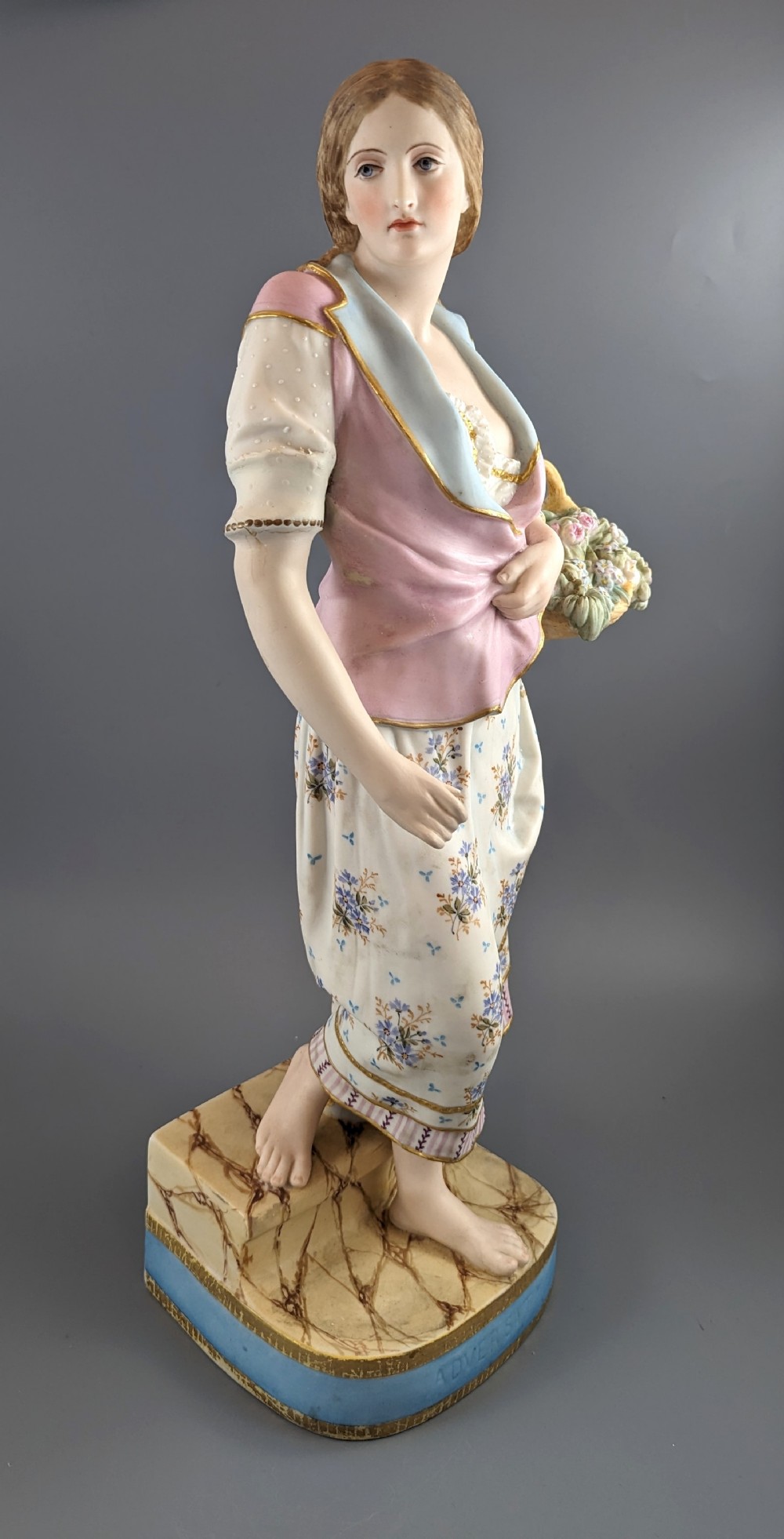 A Victorian coloured Parian porcelain figure depicting a draped female figure holding a basket of flowers on a faux-marble painted base.