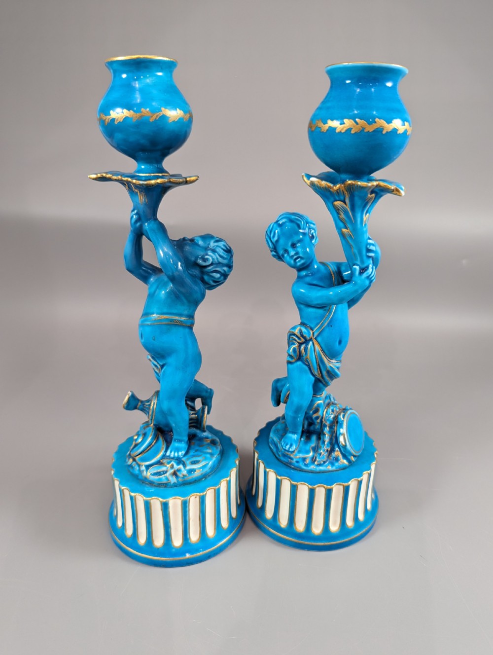 A pair of turquoise blue glazed porcelain candlesticks shaped as cherubs supporting urn sockets on fluted bases with gold trim.