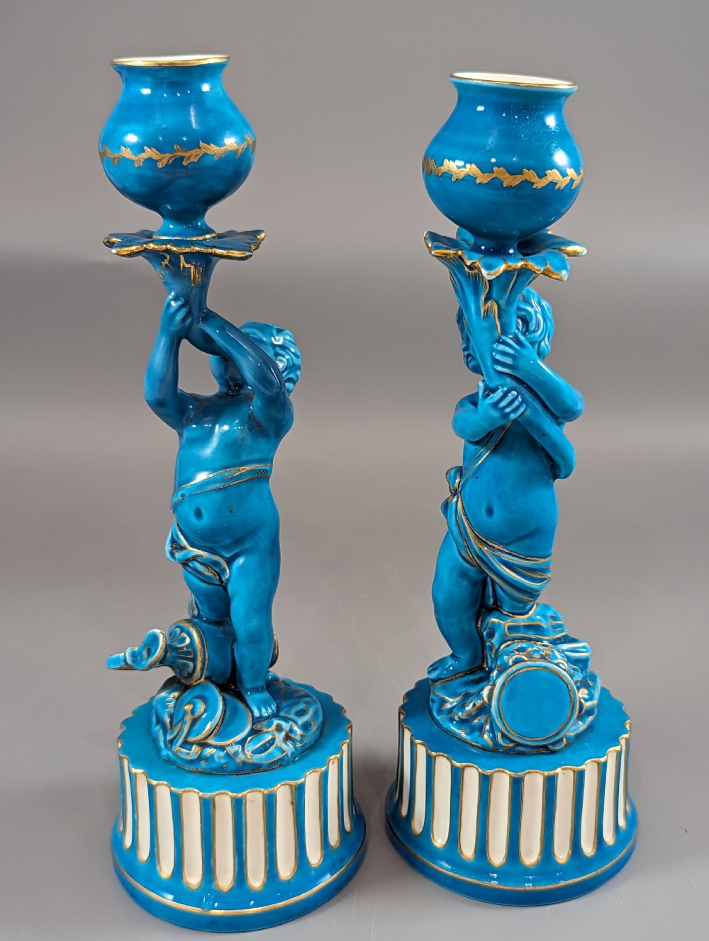 A pair of turquoise blue glazed porcelain candlesticks shaped as cherubs supporting urn sockets on fluted bases with gold trim. View XVIII