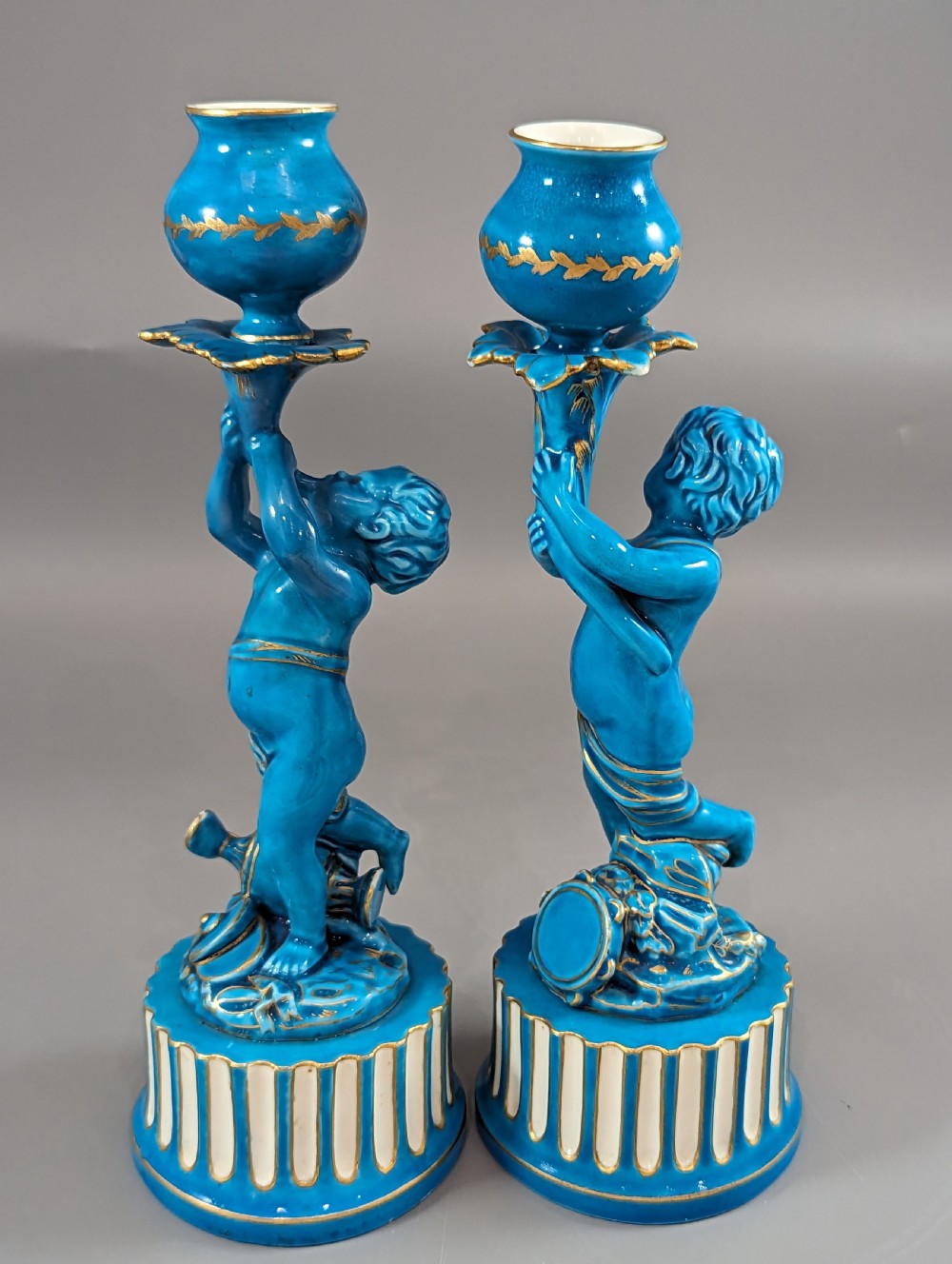 A pair of turquoise blue glazed porcelain candlesticks shaped as cherubs supporting urn sockets on fluted bases with gold trim. View XIX