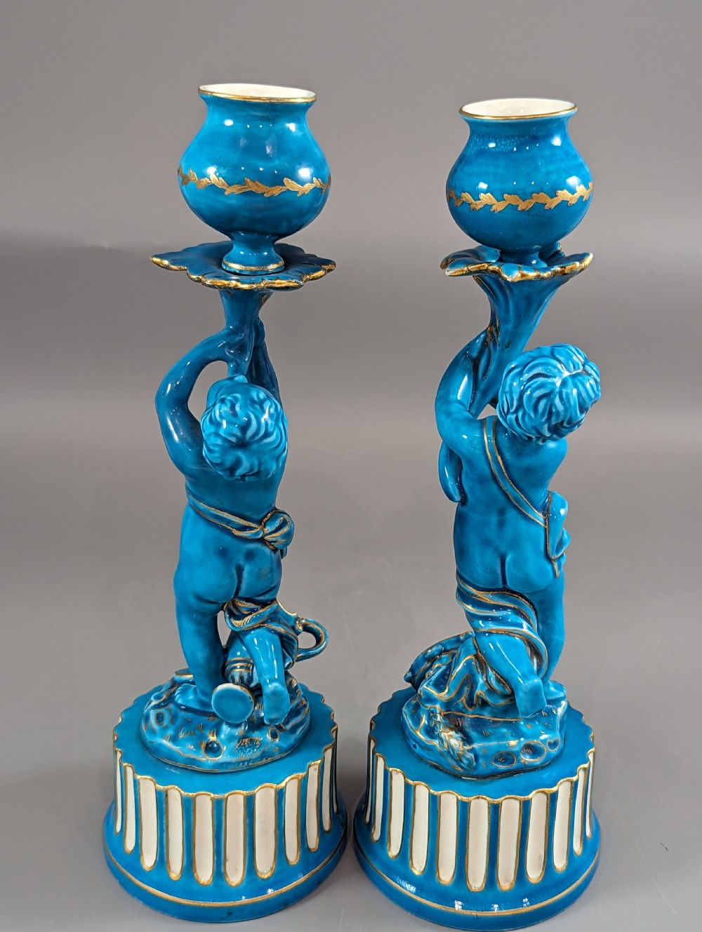 A pair of turquoise blue glazed porcelain candlesticks shaped as cherubs supporting urn sockets on fluted bases with gold trim. View XX