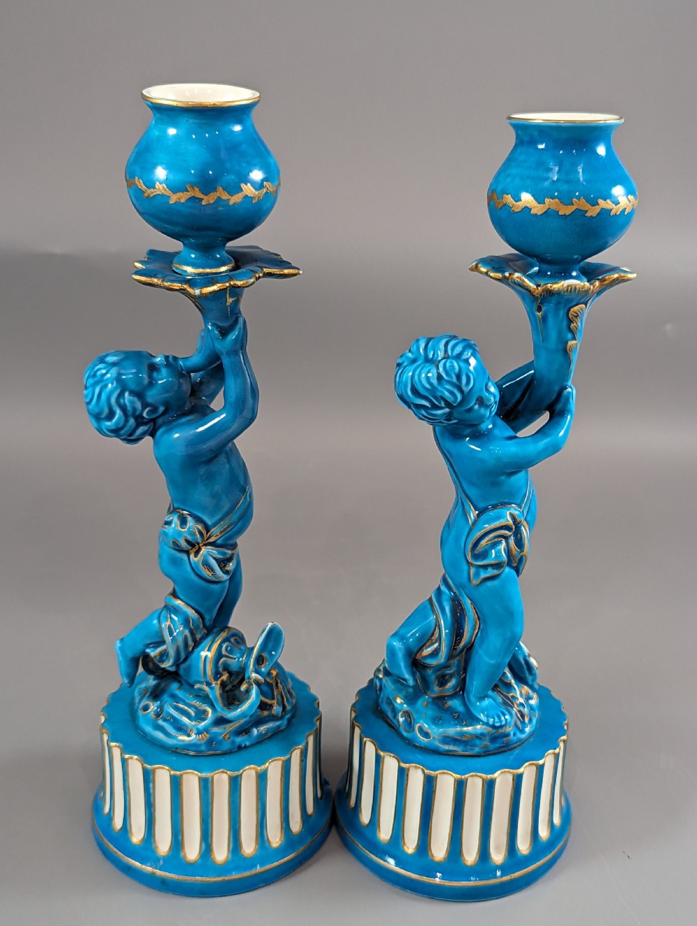 A pair of turquoise blue glazed porcelain candlesticks shaped as cherubs supporting urn sockets on fluted bases with gold trim. View XXI