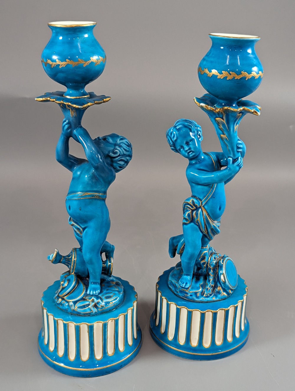 A pair of turquoise blue glazed porcelain candlesticks shaped as cherubs supporting urn sockets on fluted bases with gold trim. View XXII
