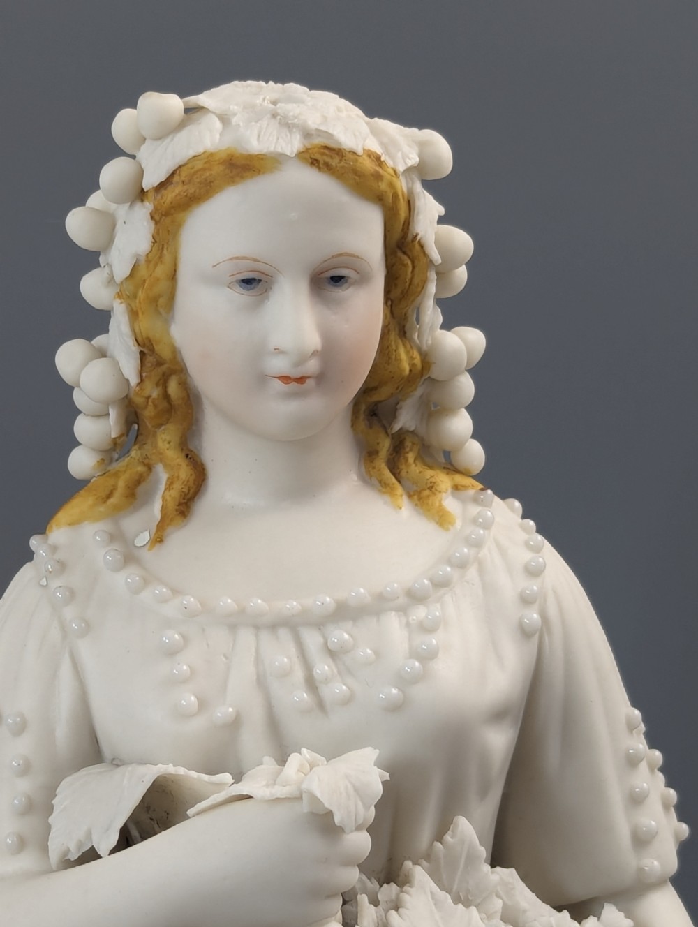 A mid-19th-century off-white Parian porcelain figurine depicting a standing lady holding an overflowing basket of grapes. View II