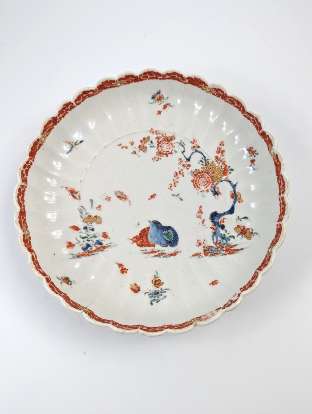 A circular fluted soft-paste porcelain dish decorated with the Kakiemon quail pattern in iron-red, blue, and gold enamels on a white ground. View II
