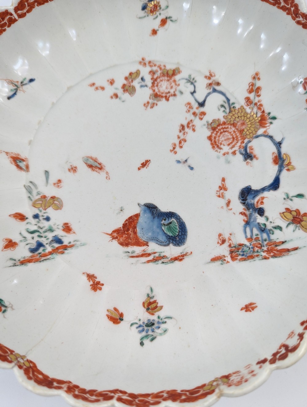 A circular fluted soft-paste porcelain dish decorated with the Kakiemon quail pattern in iron-red, blue, and gold enamels on a white ground. View III
