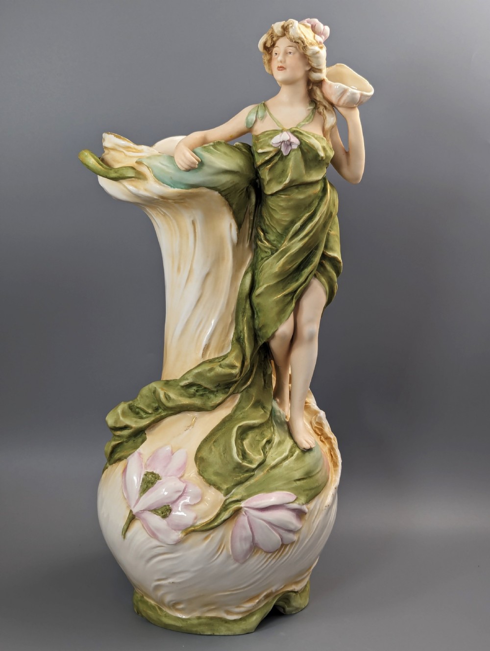 An Art Nouveau Royal Dux porcelain vase depicting a draped water nymph holding a seashell against flowing foliage in cream, olive green, and soft pink shades.