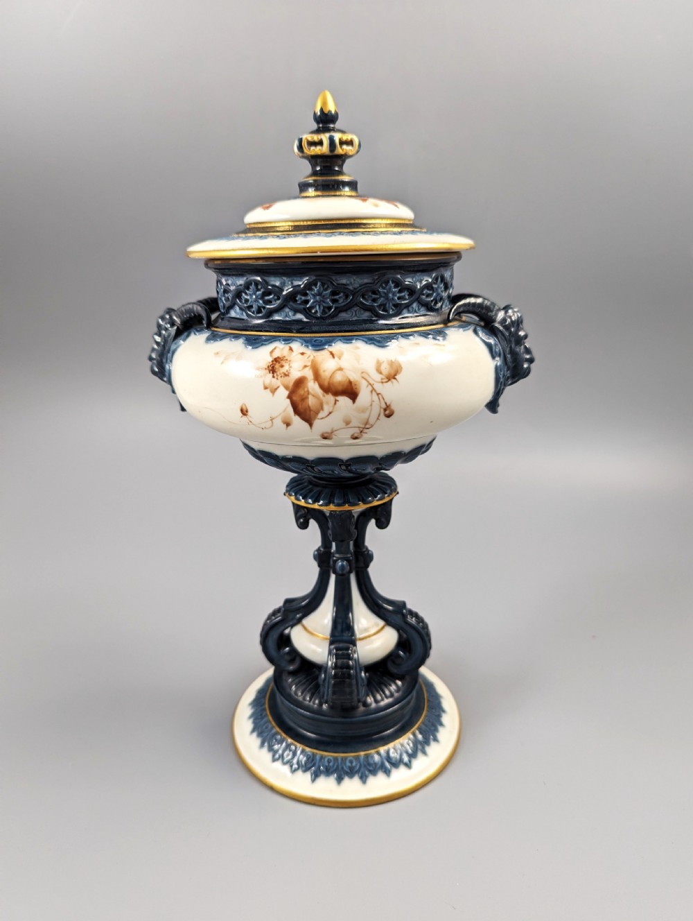 A late Victorian cream and dark blue porcelain pedestal urn decorated with hand-painted sepia floral panels, dark scroll handles, a matching lid, and gilded accents.