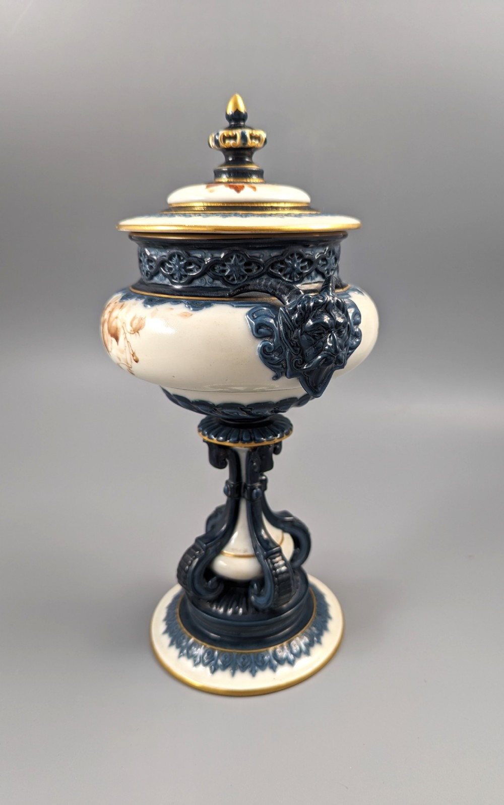 A late Victorian cream and dark blue porcelain pedestal urn decorated with hand-painted sepia floral panels, dark scroll handles, a matching lid, and gilded accents. View II