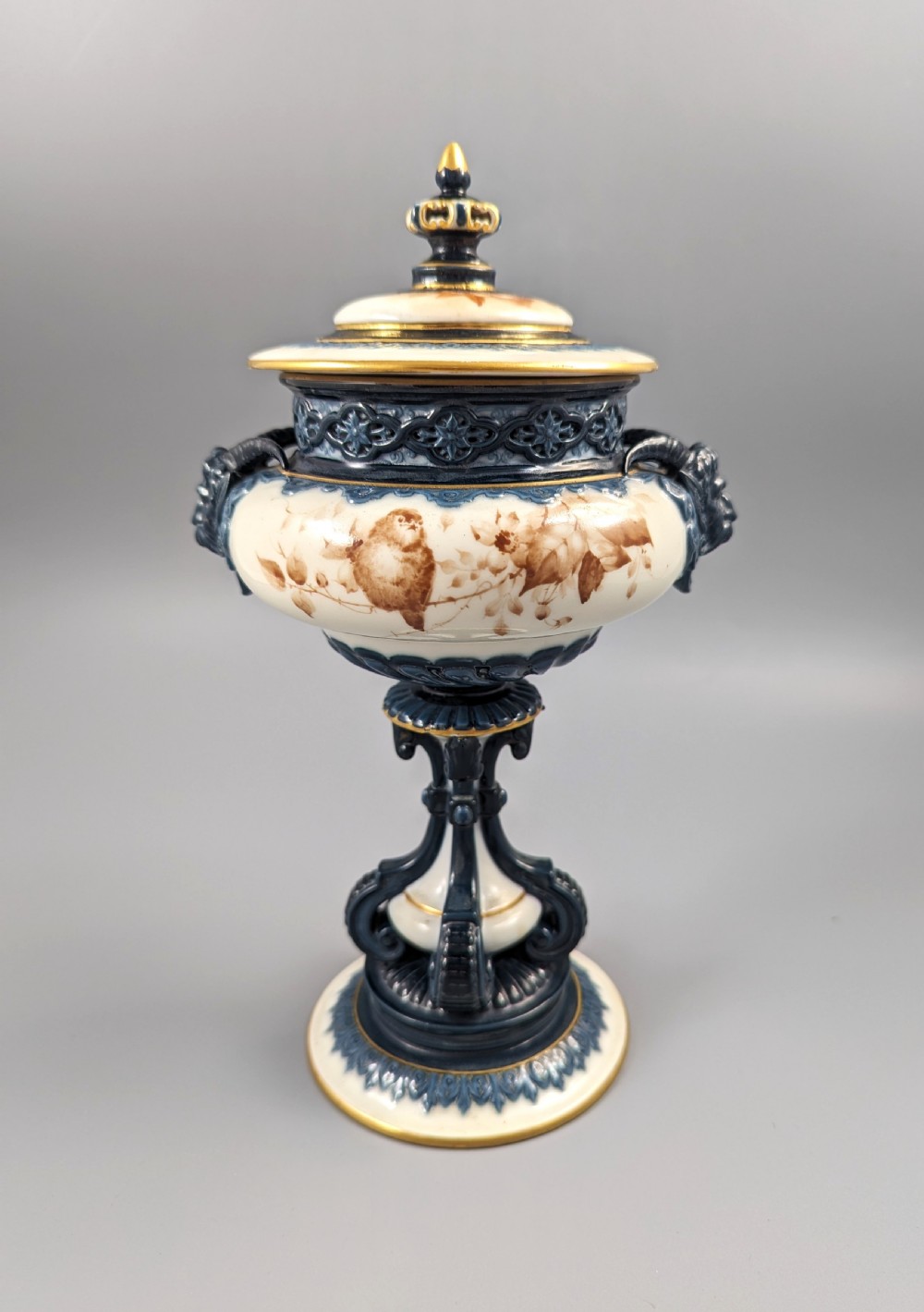 A late Victorian cream and dark blue porcelain pedestal urn decorated with hand-painted sepia floral panels, dark scroll handles, a matching lid, and gilded accents. View IV