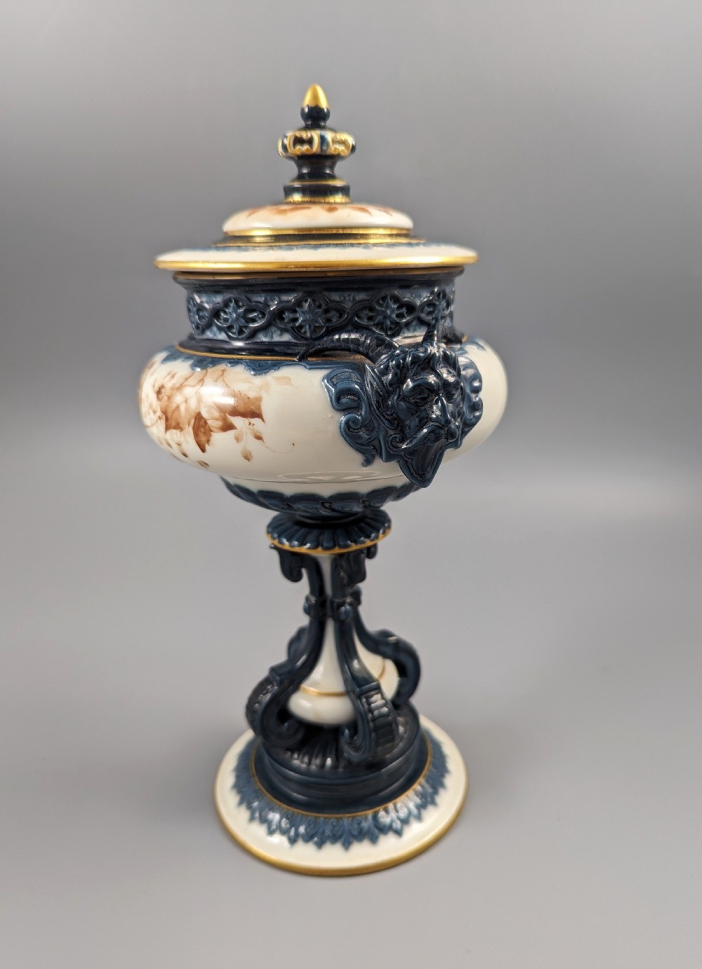 A late Victorian cream and dark blue porcelain pedestal urn decorated with hand-painted sepia floral panels, dark scroll handles, a matching lid, and gilded accents. View V