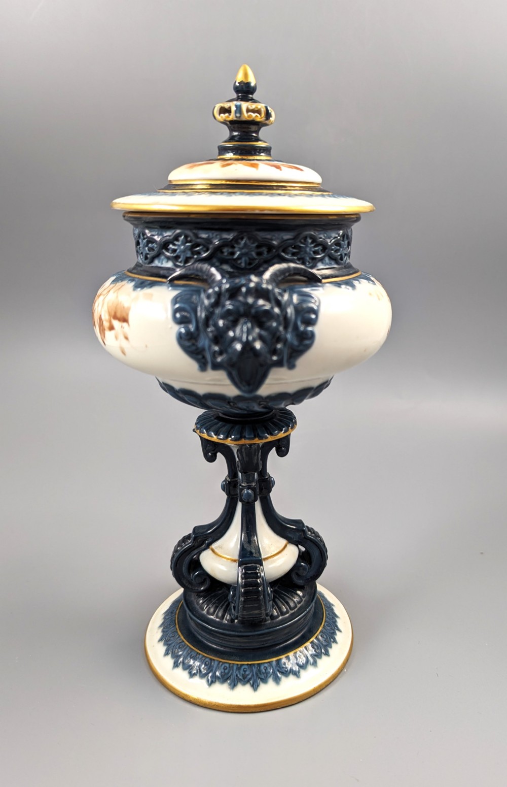 A late Victorian cream and dark blue porcelain pedestal urn decorated with hand-painted sepia floral panels, dark scroll handles, a matching lid, and gilded accents. View VI