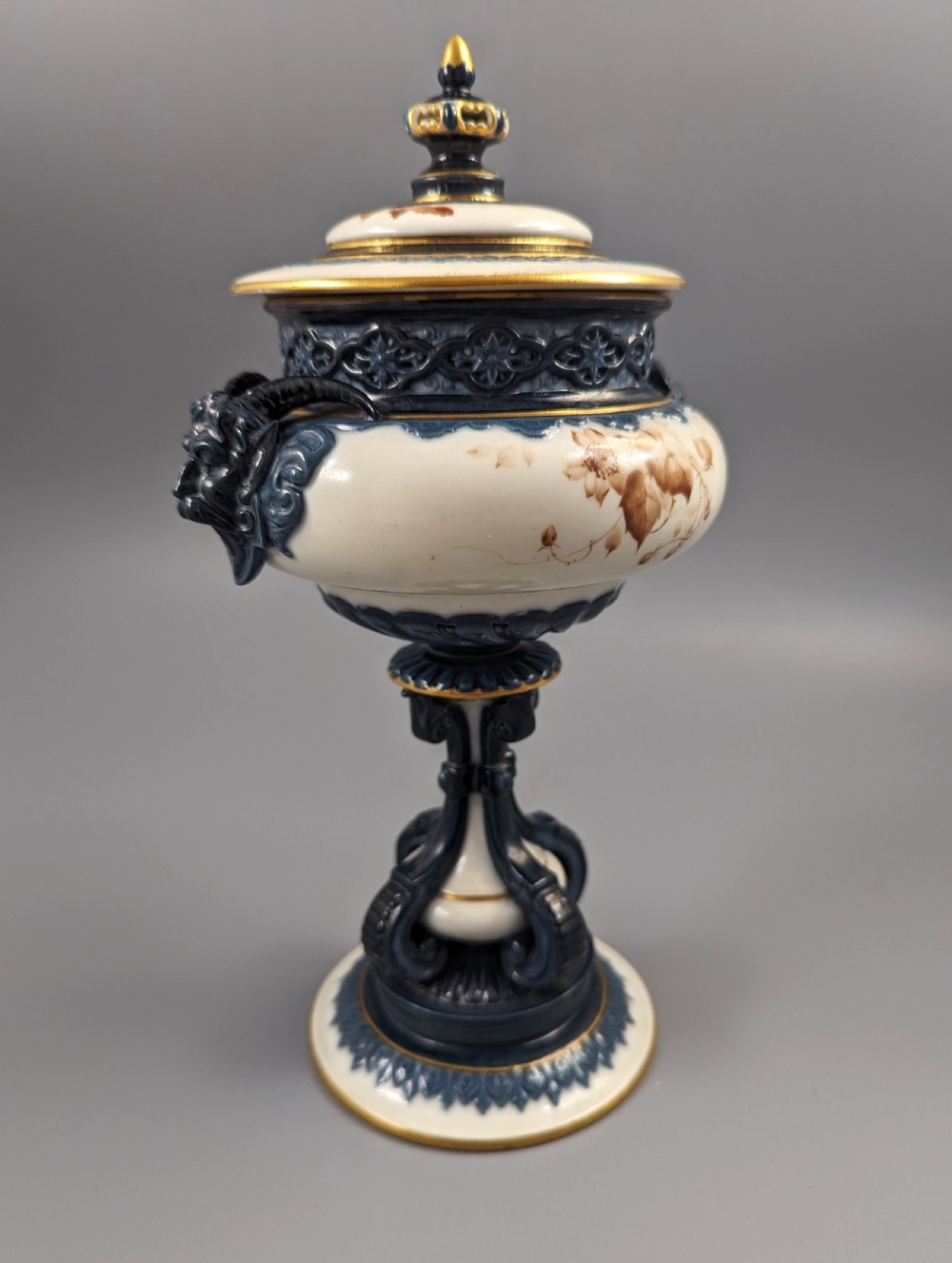 A late Victorian cream and dark blue porcelain pedestal urn decorated with hand-painted sepia floral panels, dark scroll handles, a matching lid, and gilded accents. View VII