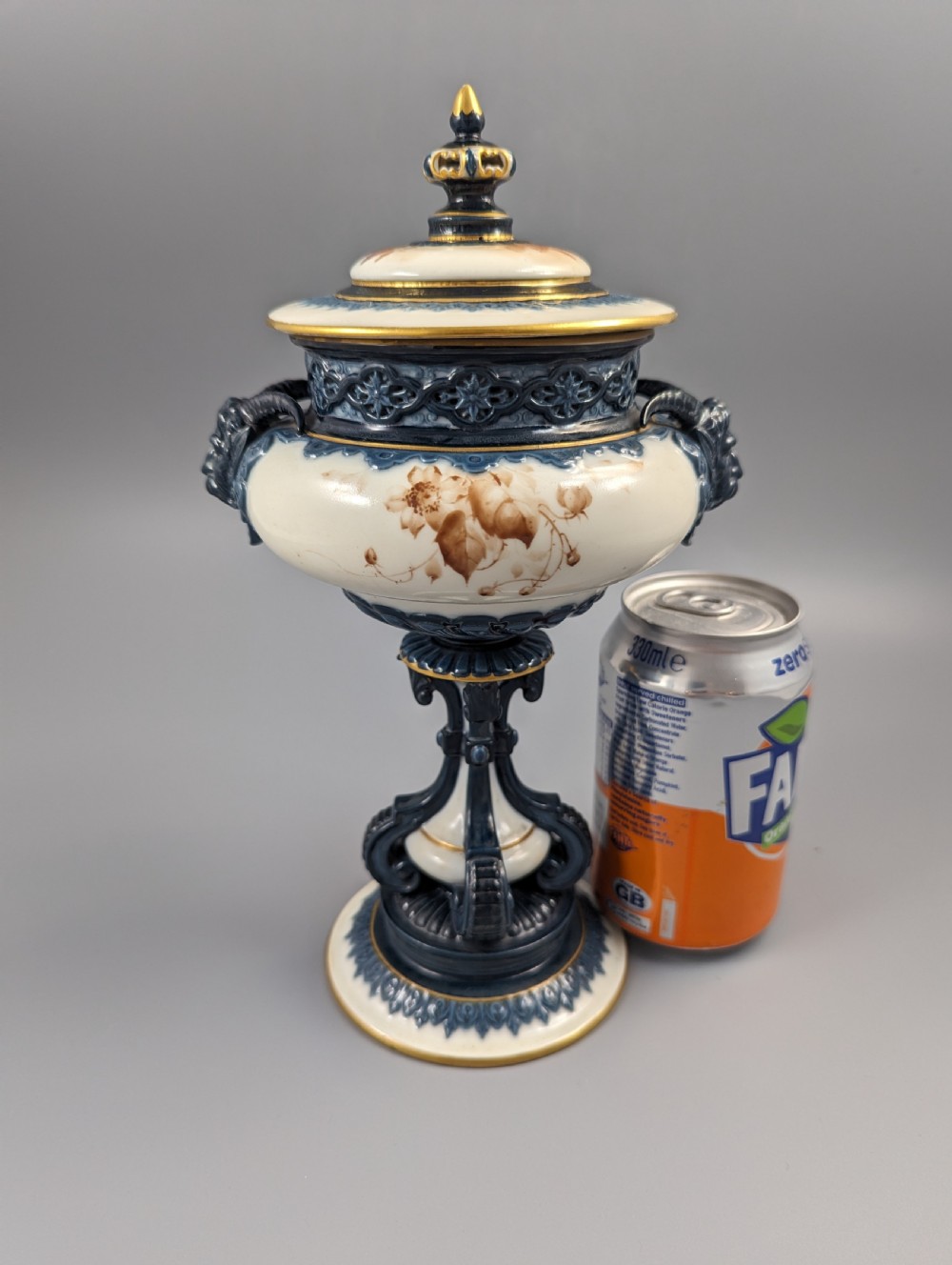 A late Victorian cream and dark blue porcelain pedestal urn decorated with hand-painted sepia floral panels, dark scroll handles, a matching lid, and gilded accents. View X