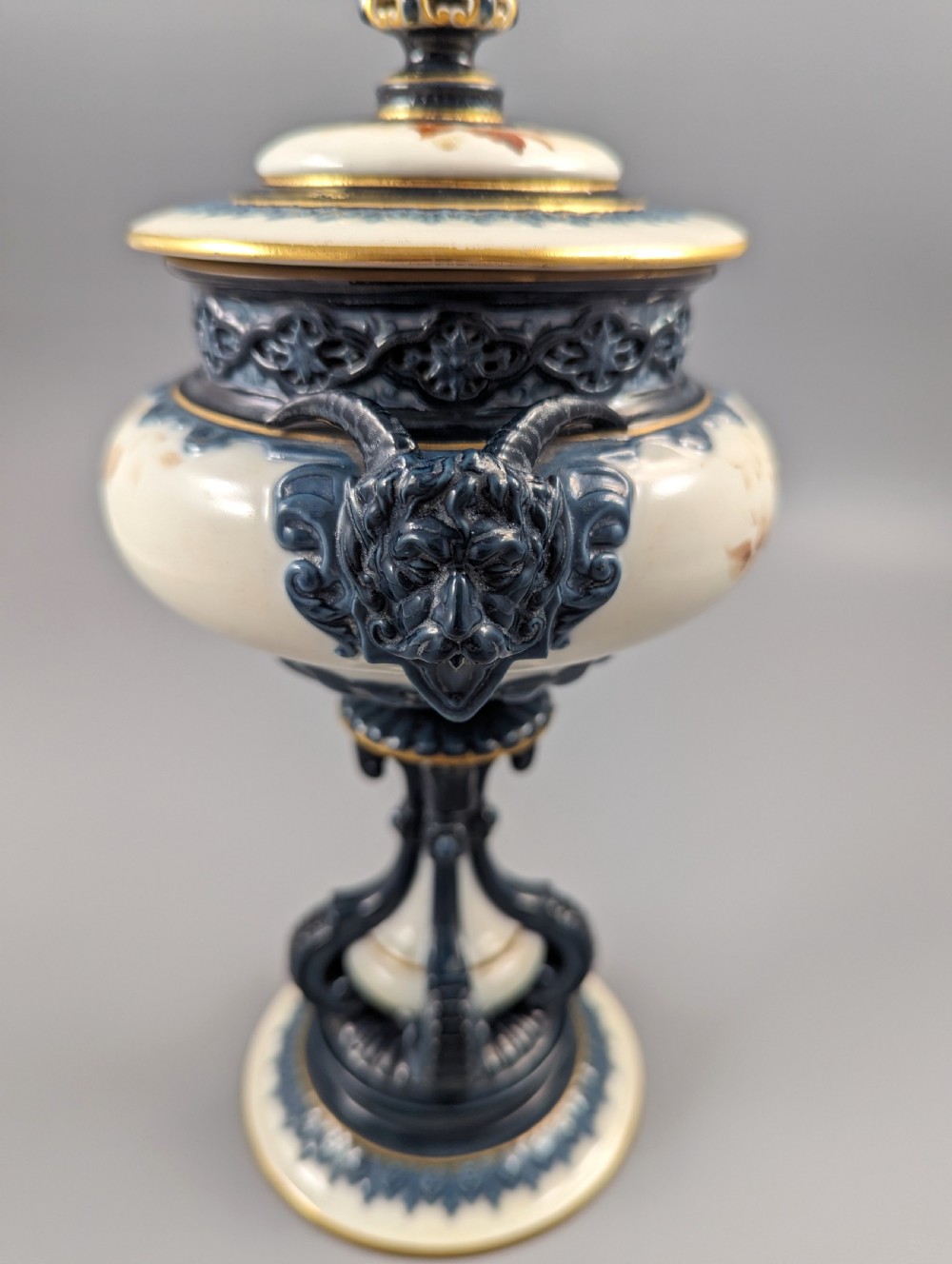 A late Victorian cream and dark blue porcelain pedestal urn decorated with hand-painted sepia floral panels, dark scroll handles, a matching lid, and gilded accents. View XV