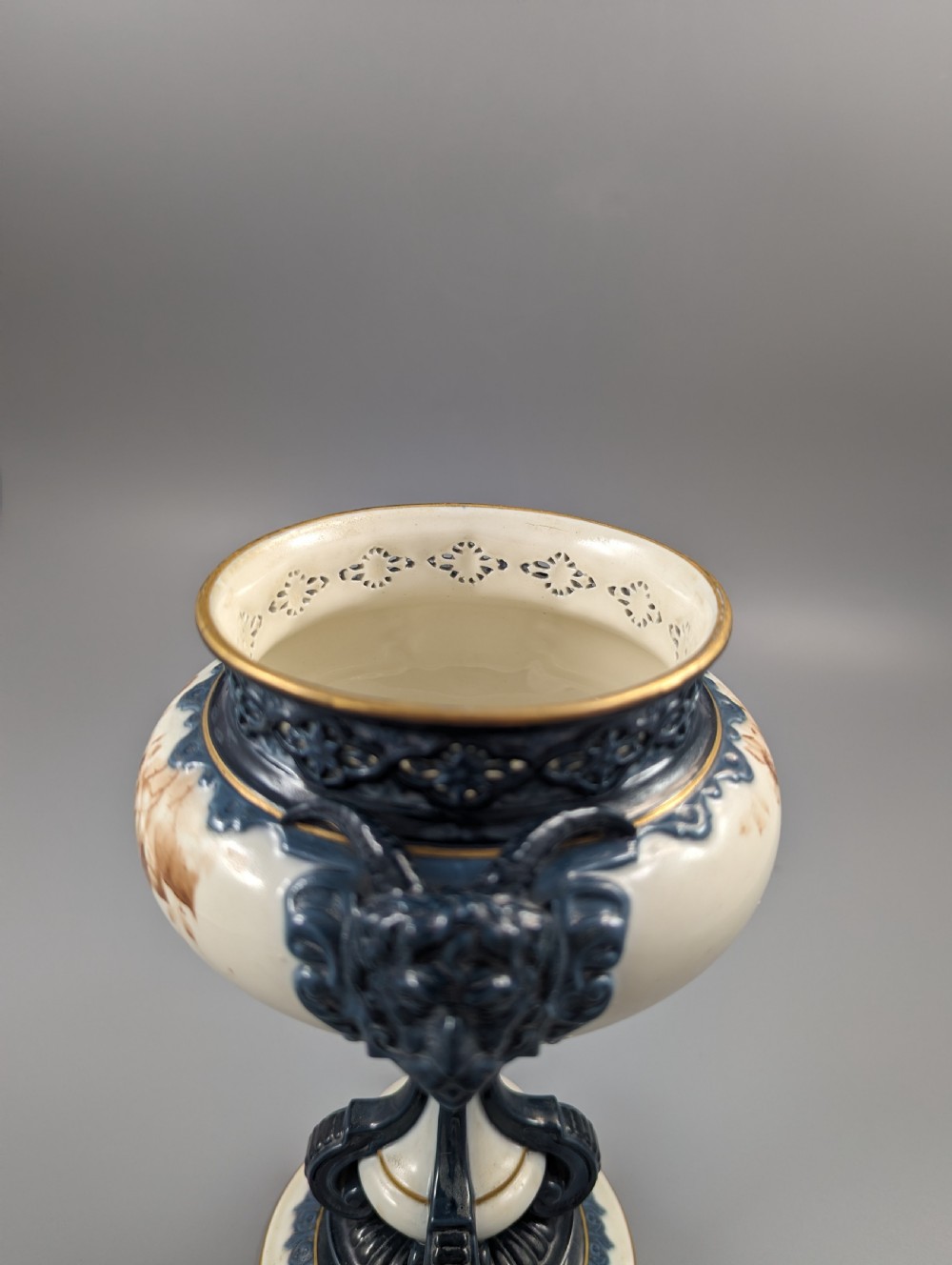 A late Victorian cream and dark blue porcelain pedestal urn decorated with hand-painted sepia floral panels, dark scroll handles, a matching lid, and gilded accents. View XVIII