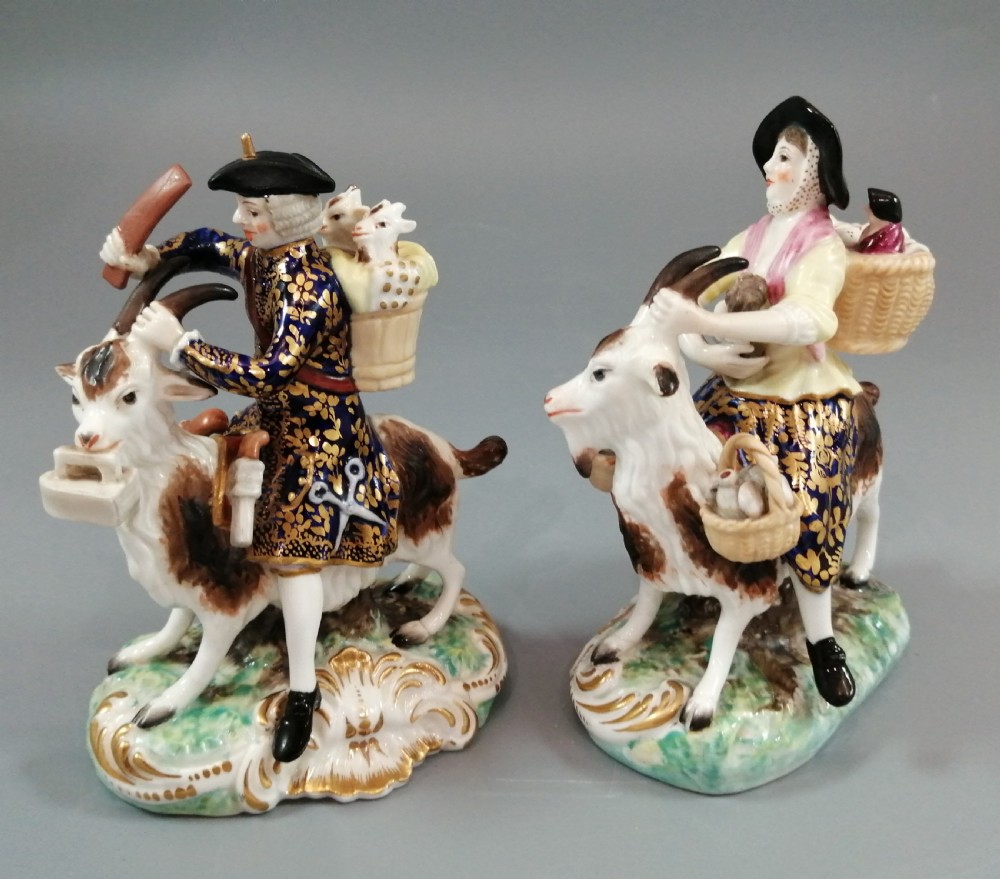A pair of painted porcelain figurines depicting a man and woman in 18th-century attire riding goats with cobalt blue gilded jackets on rococo scroll bases.