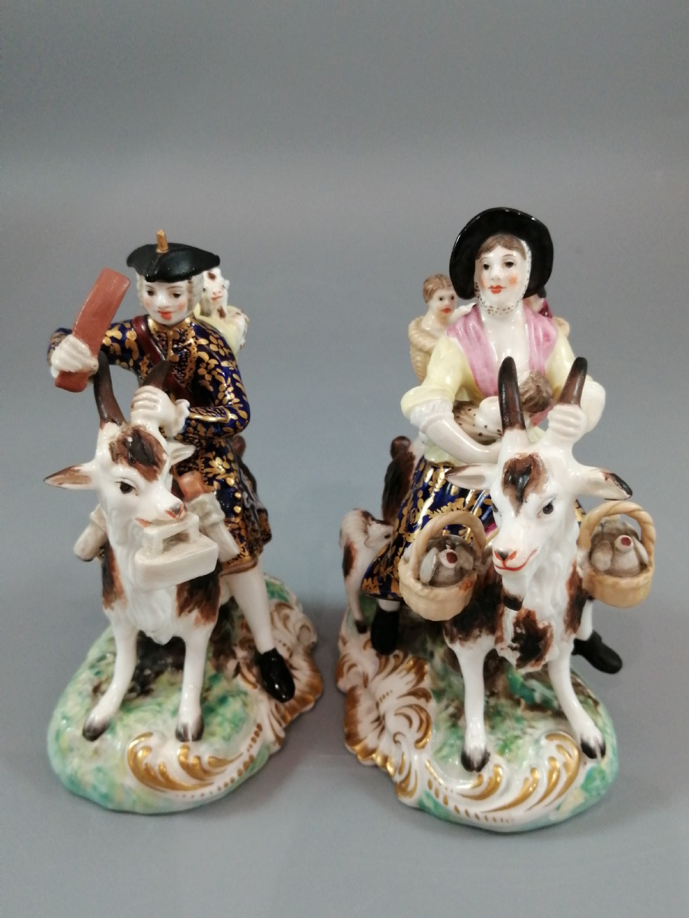 A pair of painted porcelain figurines depicting a man and woman in 18th-century attire riding goats with cobalt blue gilded jackets on rococo scroll bases. View II