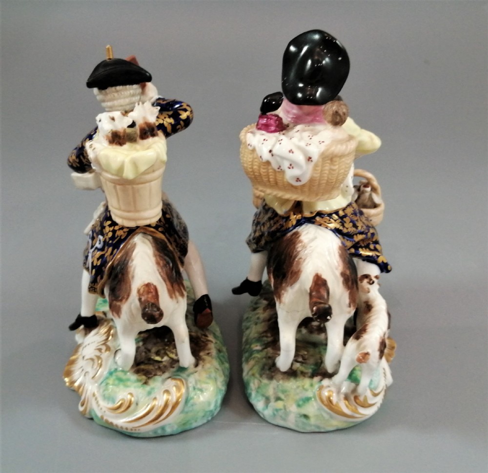 A pair of painted porcelain figurines depicting a man and woman in 18th-century attire riding goats with cobalt blue gilded jackets on rococo scroll bases. View IV