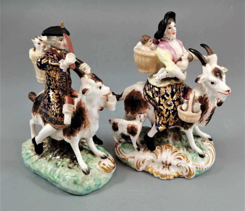 A pair of painted porcelain figurines depicting a man and woman in 18th-century attire riding goats with cobalt blue gilded jackets on rococo scroll bases. View V