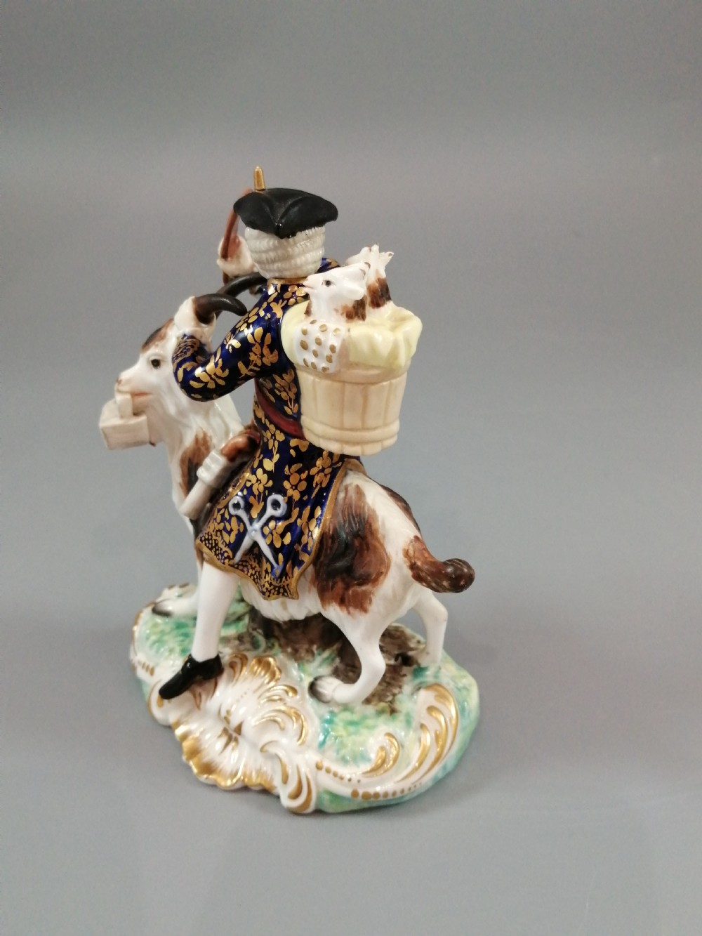 A pair of painted porcelain figurines depicting a man and woman in 18th-century attire riding goats with cobalt blue gilded jackets on rococo scroll bases. View VIII