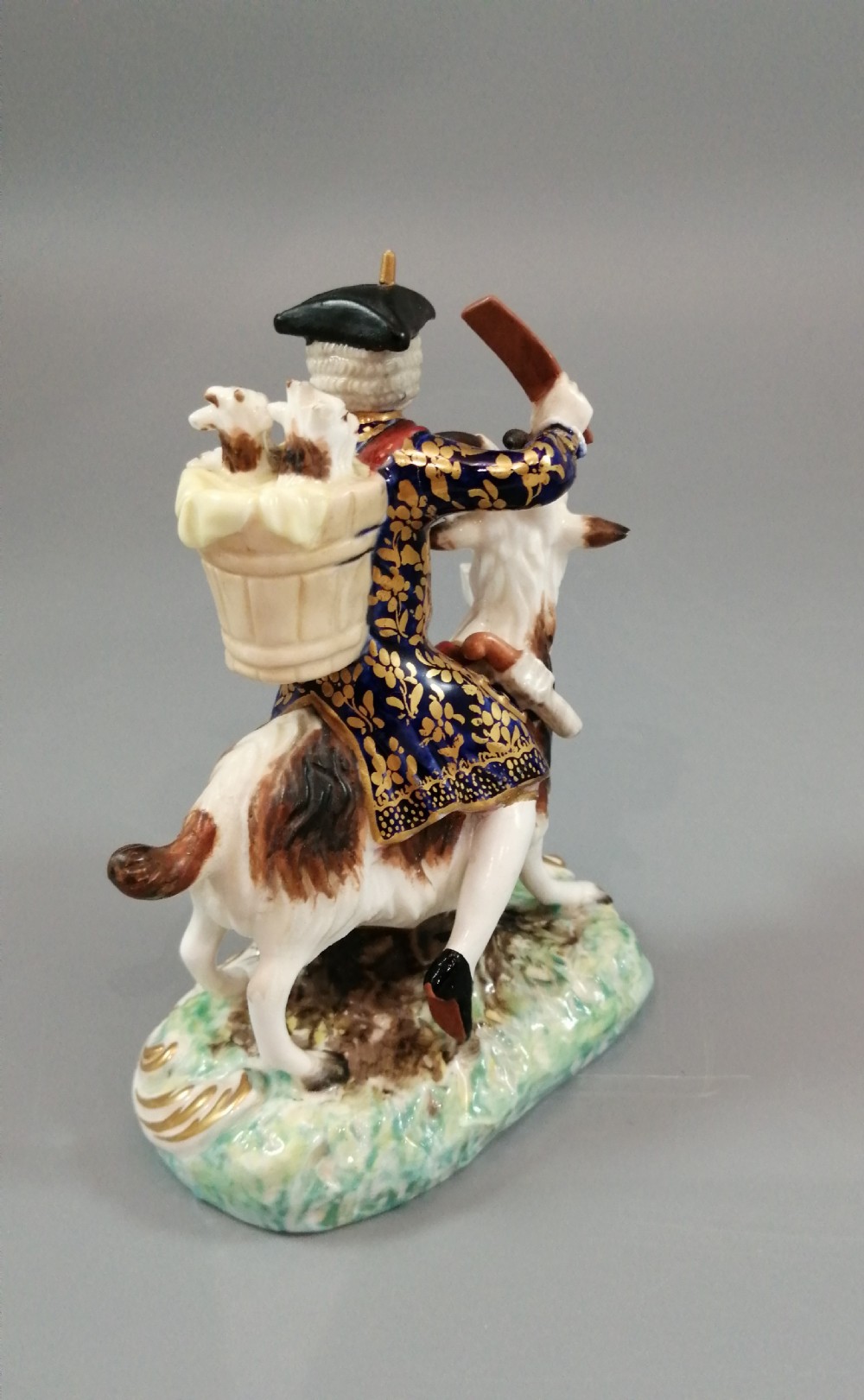 A pair of painted porcelain figurines depicting a man and woman in 18th-century attire riding goats with cobalt blue gilded jackets on rococo scroll bases. View IX