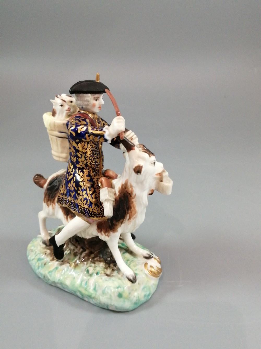 A pair of painted porcelain figurines depicting a man and woman in 18th-century attire riding goats with cobalt blue gilded jackets on rococo scroll bases. View X