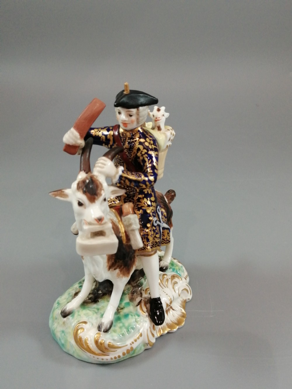 A pair of painted porcelain figurines depicting a man and woman in 18th-century attire riding goats with cobalt blue gilded jackets on rococo scroll bases. View XI