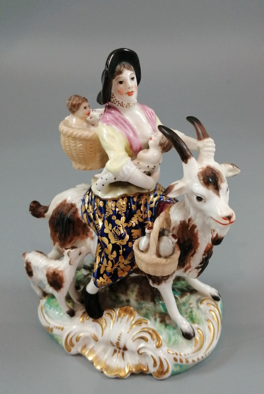 A pair of painted porcelain figurines depicting a man and woman in 18th-century attire riding goats with cobalt blue gilded jackets on rococo scroll bases. View XIII