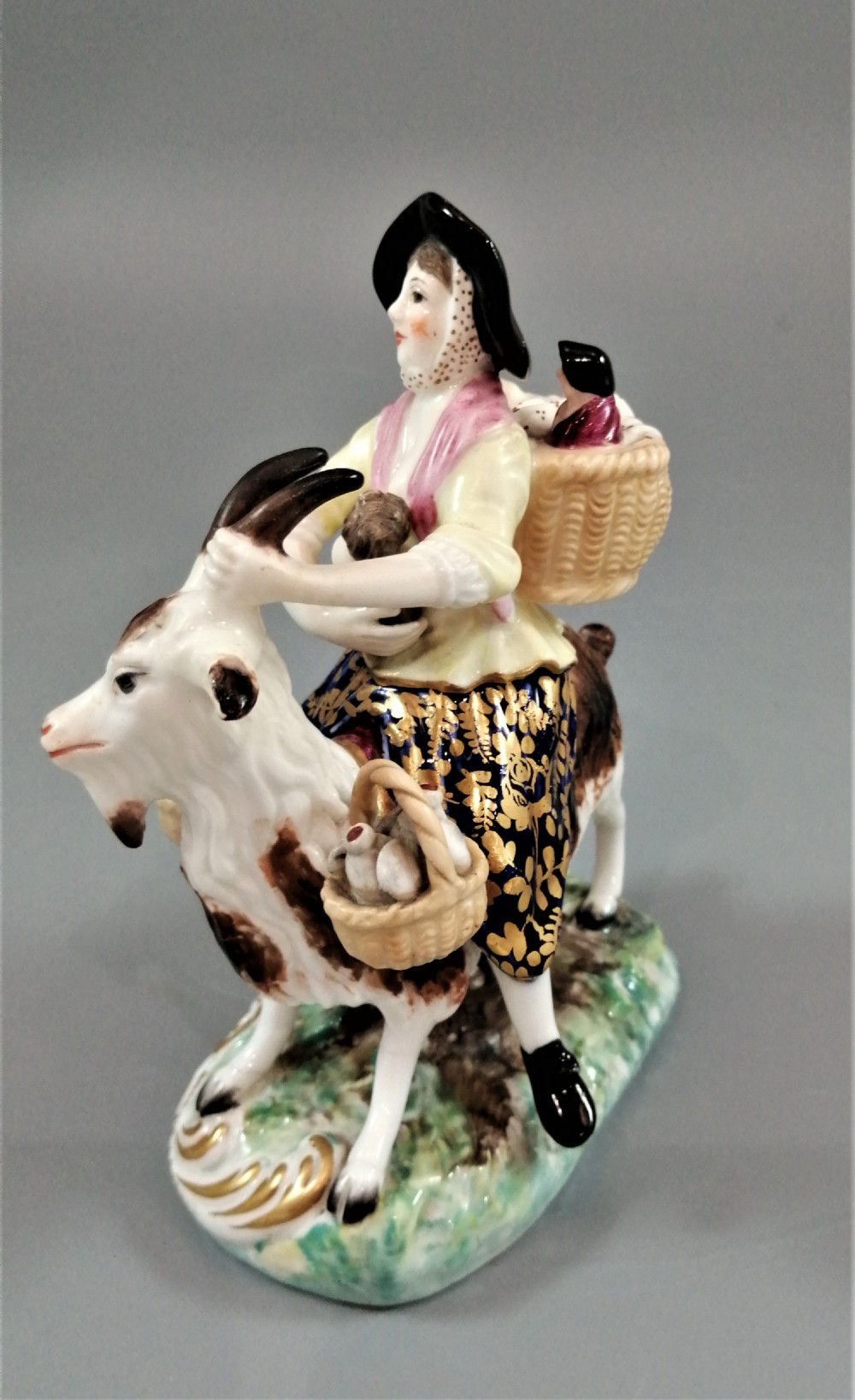 A pair of painted porcelain figurines depicting a man and woman in 18th-century attire riding goats with cobalt blue gilded jackets on rococo scroll bases. View XV