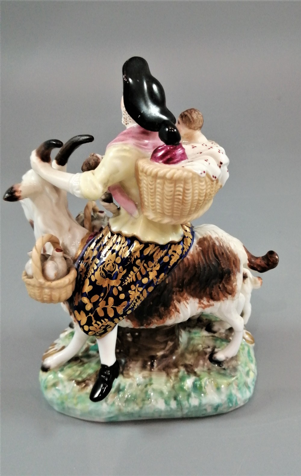 A pair of painted porcelain figurines depicting a man and woman in 18th-century attire riding goats with cobalt blue gilded jackets on rococo scroll bases. View XVI