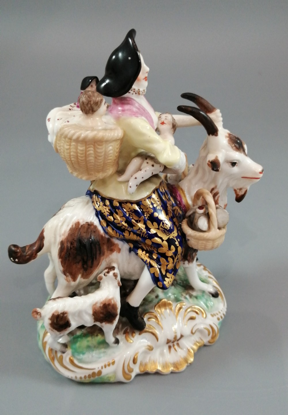 A pair of painted porcelain figurines depicting a man and woman in 18th-century attire riding goats with cobalt blue gilded jackets on rococo scroll bases. View XVIII