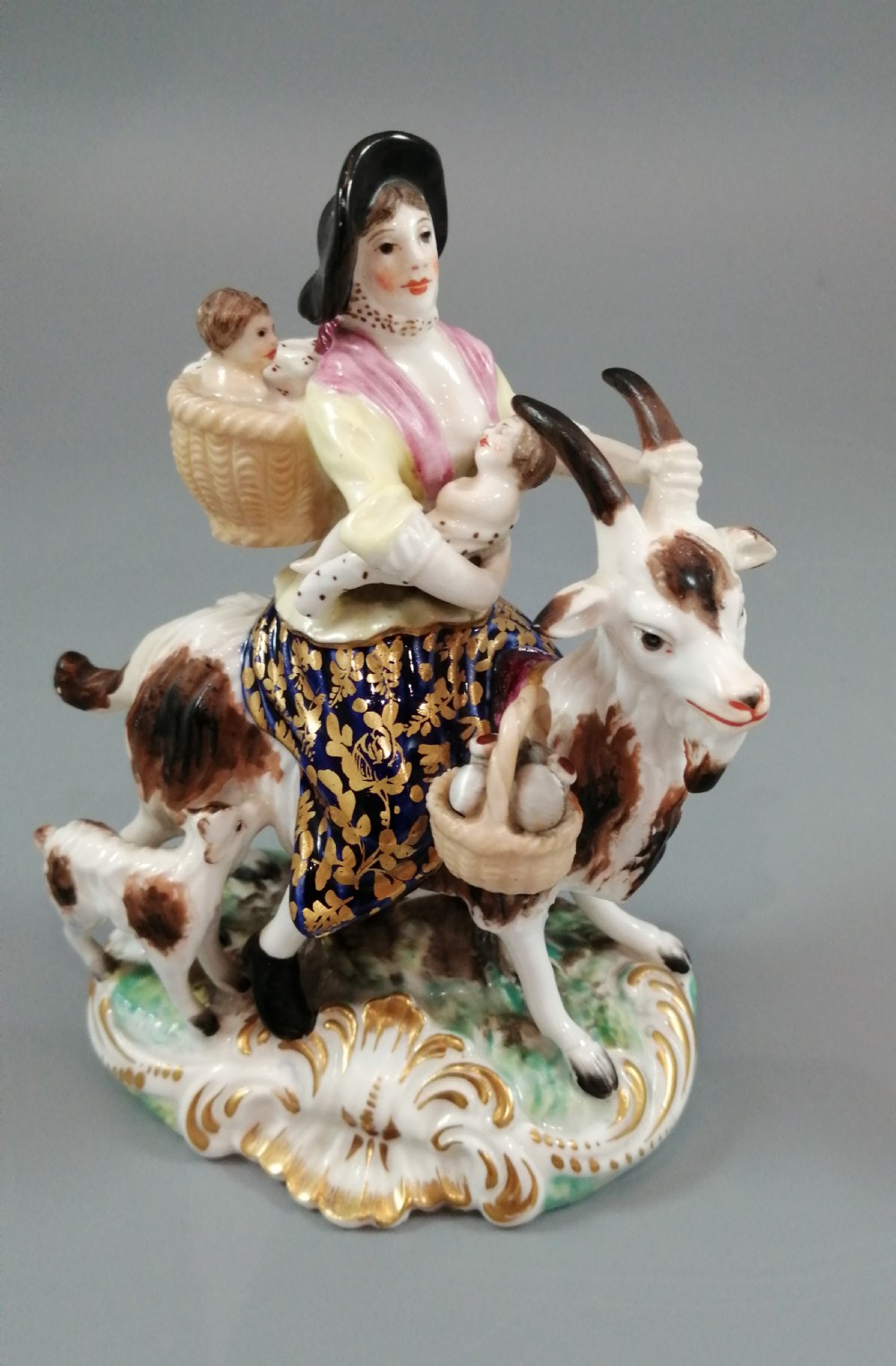 A pair of painted porcelain figurines depicting a man and woman in 18th-century attire riding goats with cobalt blue gilded jackets on rococo scroll bases. View XIX