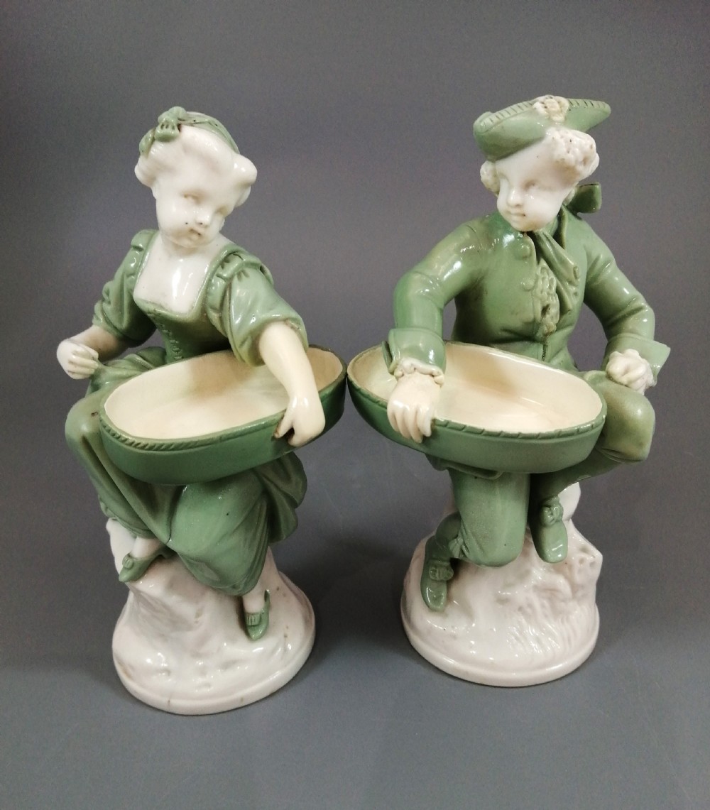A pair of Victorian Minton celadon green and white porcelain figurines depicting a seated man and woman holding oval dishes on circular bases.