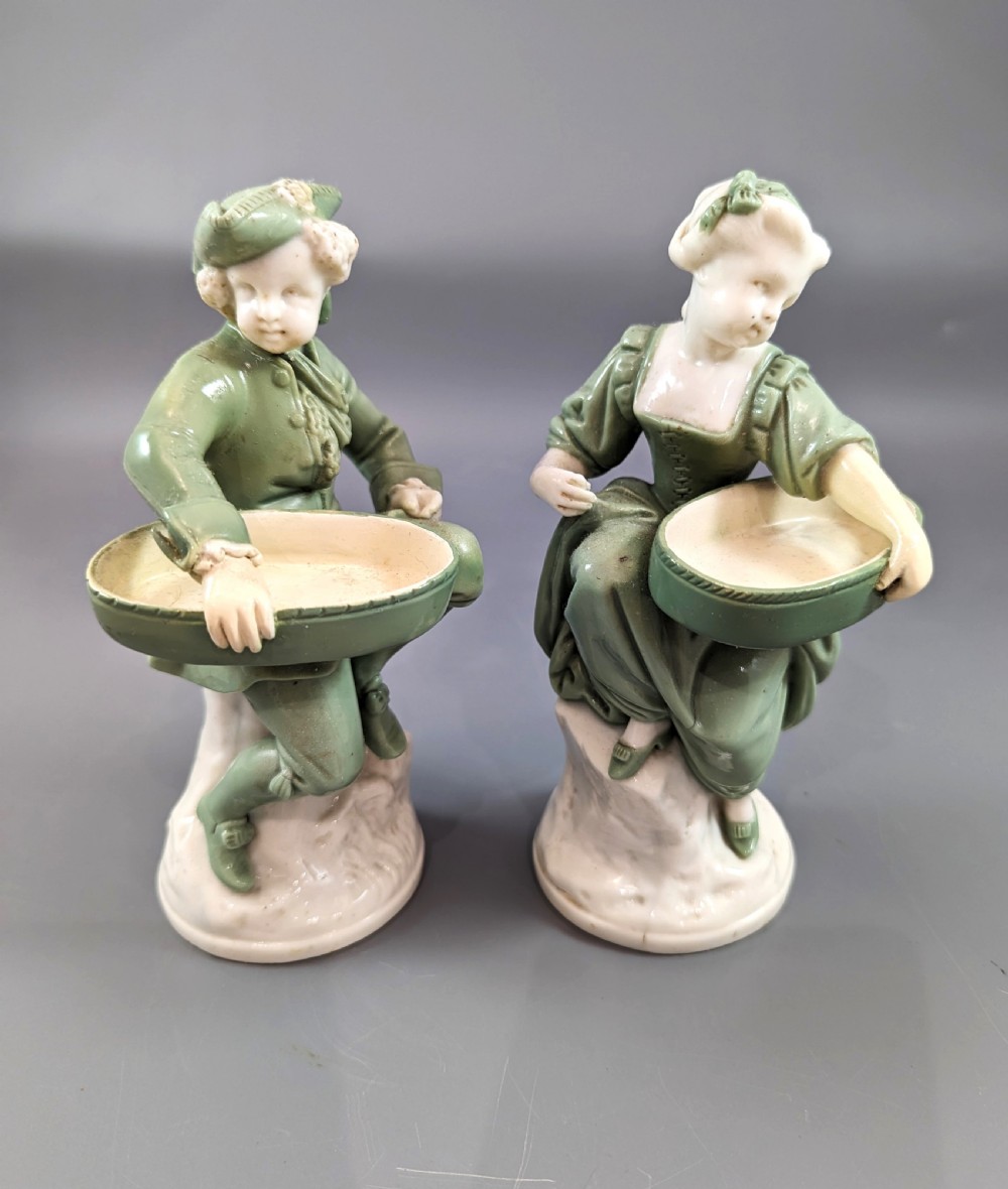 A pair of Victorian Minton celadon green and white porcelain figurines depicting a seated man and woman holding oval dishes on circular bases. View II