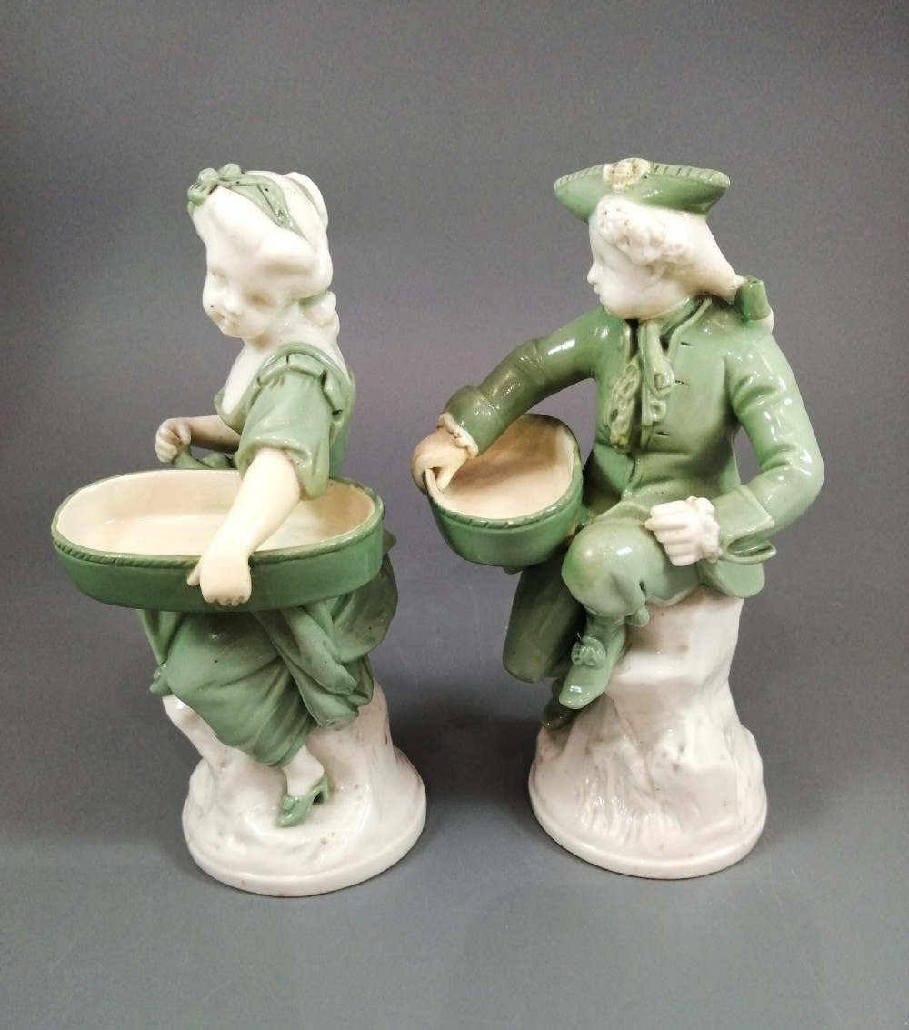 A pair of Victorian Minton celadon green and white porcelain figurines depicting a seated man and woman holding oval dishes on circular bases. View III