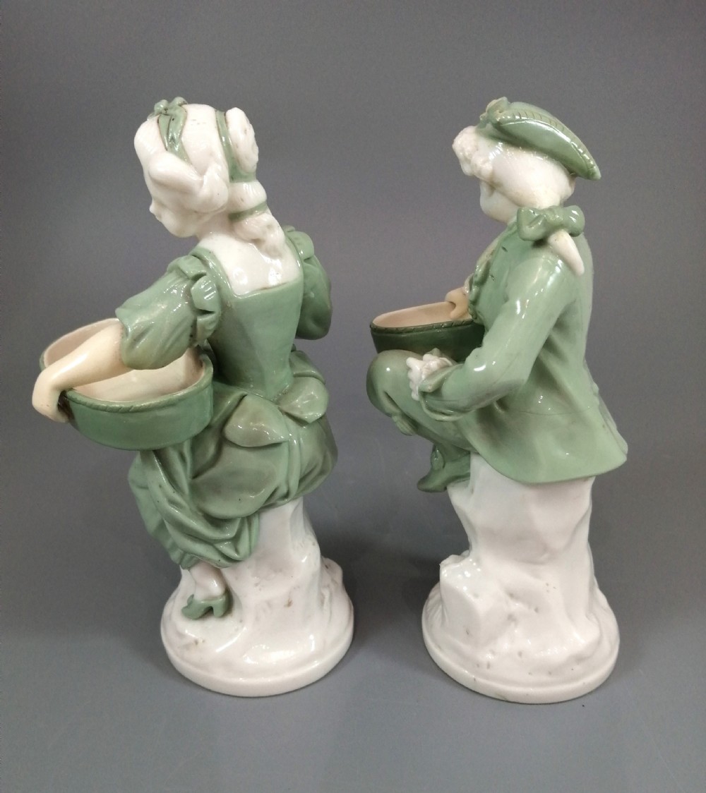 A pair of Victorian Minton celadon green and white porcelain figurines depicting a seated man and woman holding oval dishes on circular bases. View IV