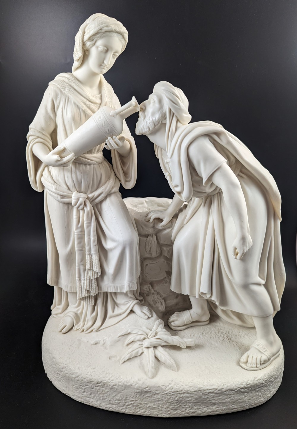 An off-white Parian porcelain sculpture depicting Rebekah offering water from an amphora to a kneeling man beside a textured stone well.
