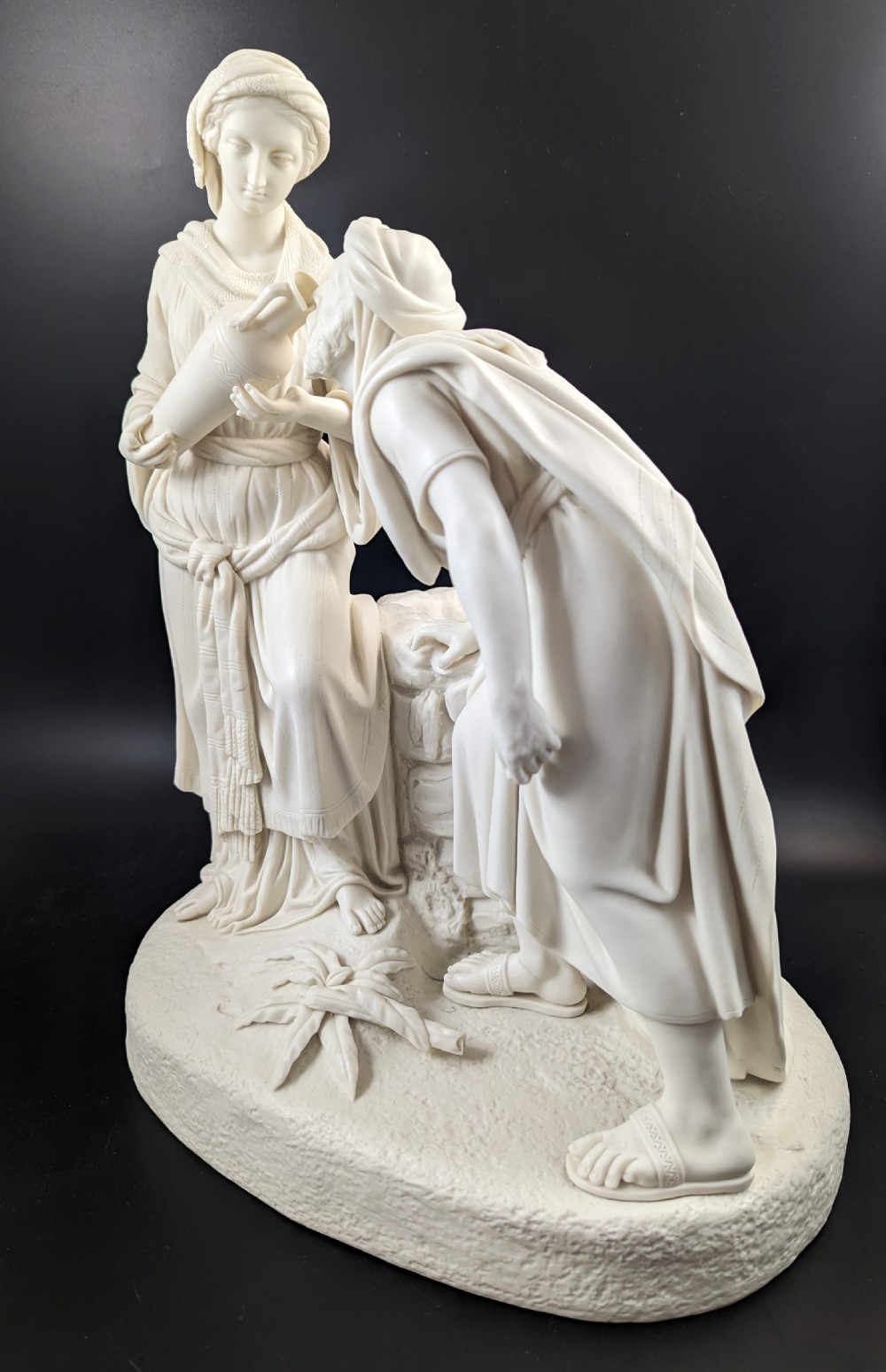 An off-white Parian porcelain sculpture depicting Rebekah offering water from an amphora to a kneeling man beside a textured stone well. View II