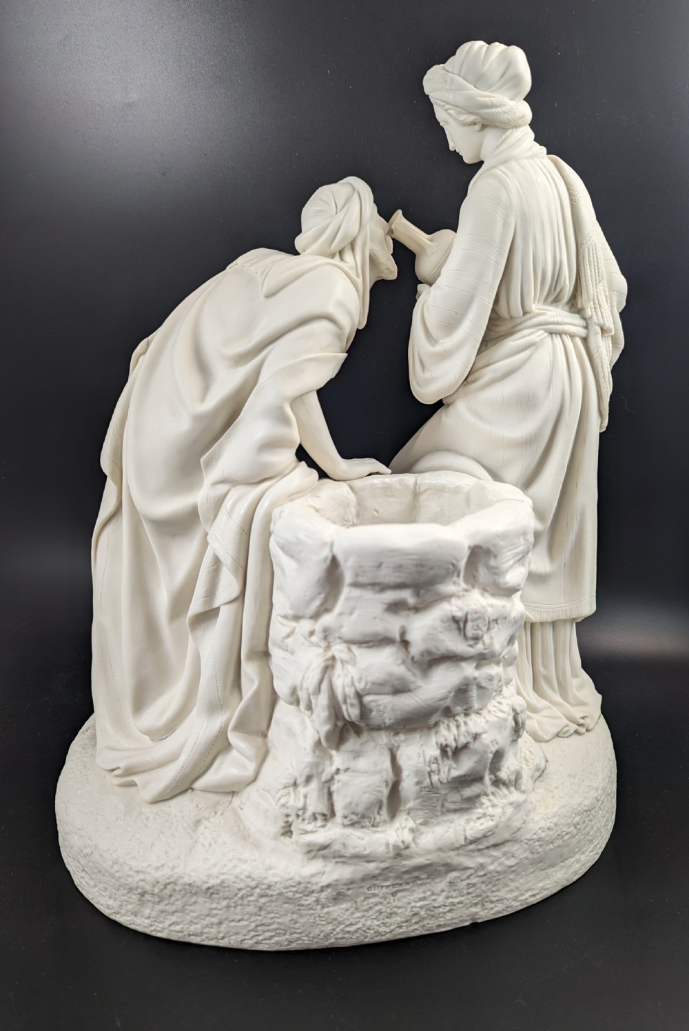 An off-white Parian porcelain sculpture depicting Rebekah offering water from an amphora to a kneeling man beside a textured stone well. View IV