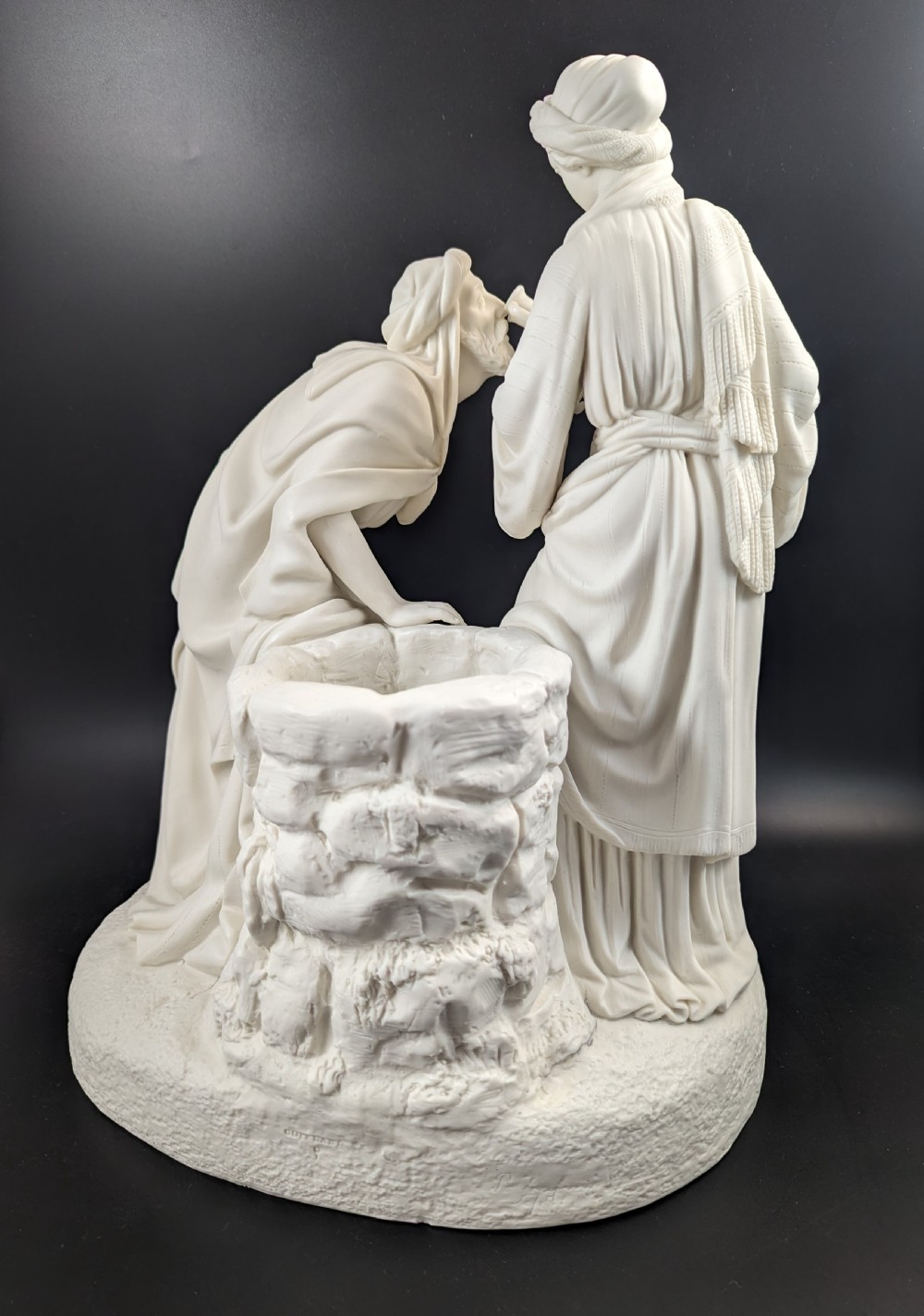 An off-white Parian porcelain sculpture depicting Rebekah offering water from an amphora to a kneeling man beside a textured stone well. View V