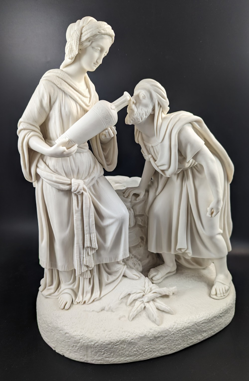 An off-white Parian porcelain sculpture depicting Rebekah offering water from an amphora to a kneeling man beside a textured stone well. View IX
