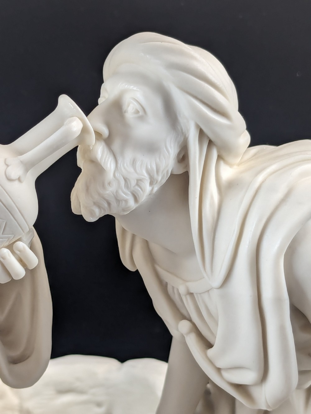 An off-white Parian porcelain sculpture depicting Rebekah offering water from an amphora to a kneeling man beside a textured stone well. View X