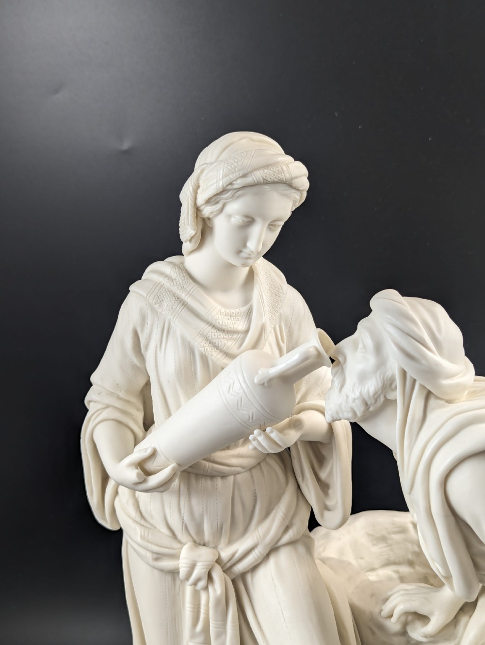 An off-white Parian porcelain sculpture depicting Rebekah offering water from an amphora to a kneeling man beside a textured stone well. View XII