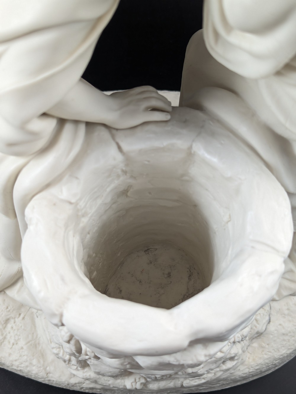An off-white Parian porcelain sculpture depicting Rebekah offering water from an amphora to a kneeling man beside a textured stone well. View XIII