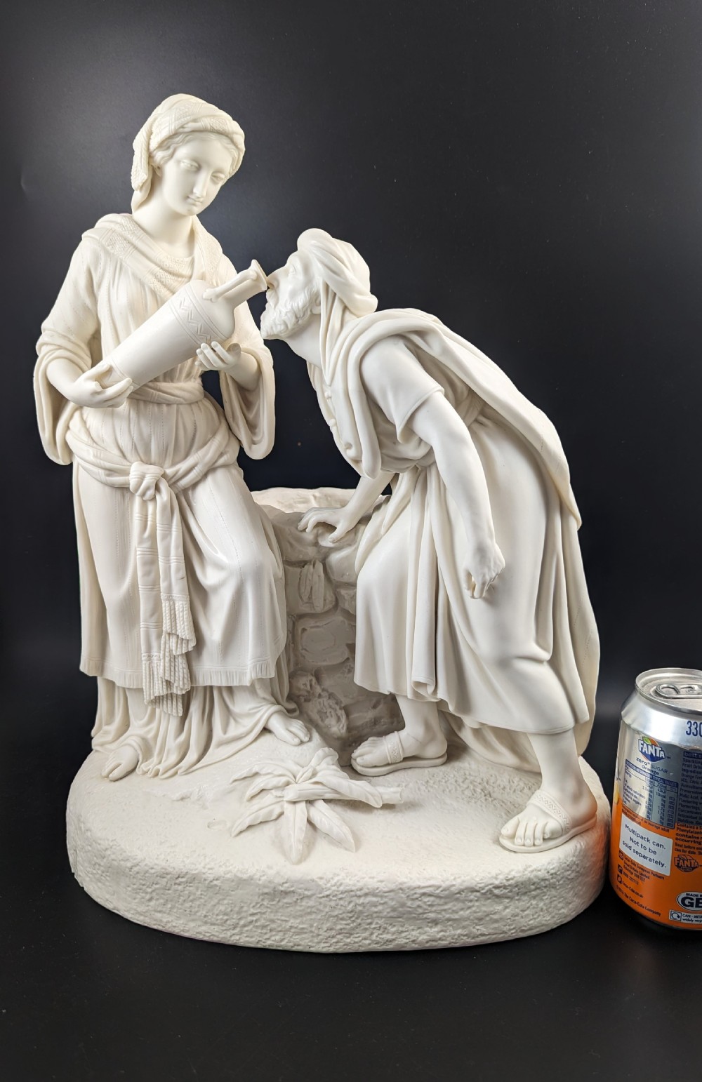 An off-white Parian porcelain sculpture depicting Rebekah offering water from an amphora to a kneeling man beside a textured stone well. View XVII